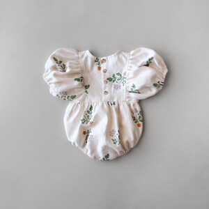 May include: A white baby onesie with a floral print of green leaves and orange flowers. The onesie has puffed sleeves and a button closure at the back.