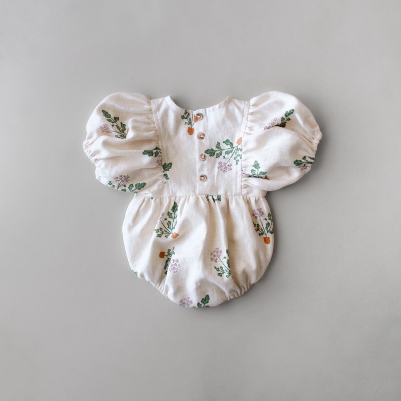 May include: A white baby onesie with a floral print of green leaves and orange flowers. The onesie has puffed sleeves and a button closure at the back.