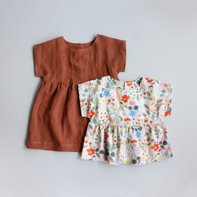 May include: Two girls' dresses. One is a solid rust-colored linen dress with short sleeves. The other is a white dress with a floral print featuring red, blue, and yellow flowers. Both dresses have a round neckline.