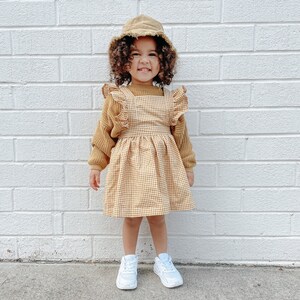 May include: A young girl wearing a yellow and white gingham dress with a ruffled neckline and sleeves. She is also wearing a brown bucket hat and white sneakers.