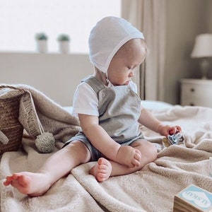 May include: A baby wearing a white bonnet and a gray overall romper sits on a light brown blanket. The baby is playing with a toy.