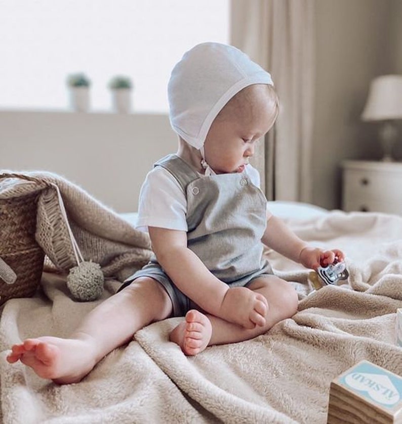 May include: A baby wearing a white bonnet and a gray overall romper sits on a light brown blanket. The baby is playing with a toy.