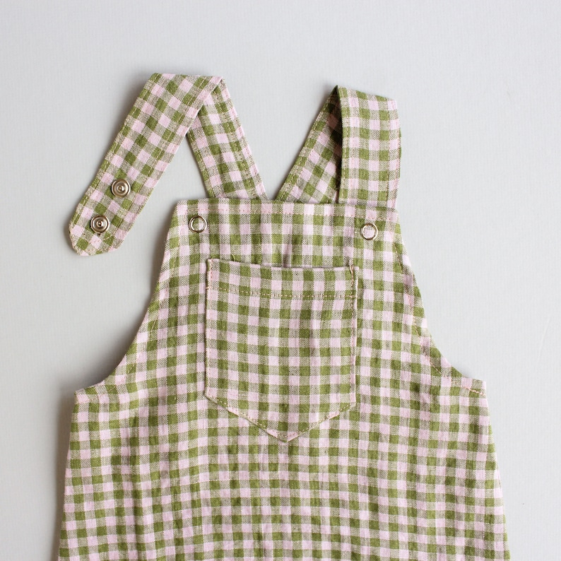 May include: Green and pink gingham overall romper with a single pocket and adjustable straps. The straps have metal button closures.