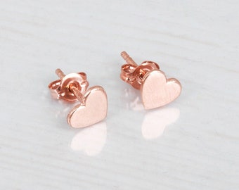 Puede incluir: Un par de pendientes de botón de oro rosa con forma de pequeños corazones. Cada pendiente presenta una superficie lisa y pulida con un mecanismo de cierre de presión estándar, mostrando un tono dorado rosado metálico y cálido.