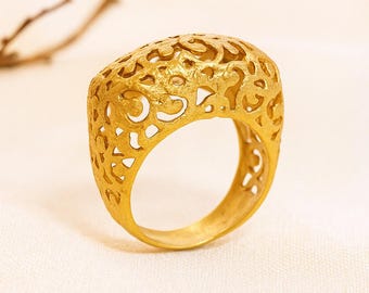 May include: A gold ring featuring an openwork design. The ring has a rectangular shape with rounded edges and a hollowed-out pattern. The intricate design allows light to pass through, creating a delicate aesthetic. The ring is set against a neutral background.