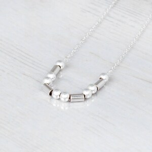 Puede incluir: Un collar de plata con una cadena delicada y un dise&ntilde;o sencillo con peque&ntilde;as cuentas de plata y separadores cil&iacute;ndricos.
