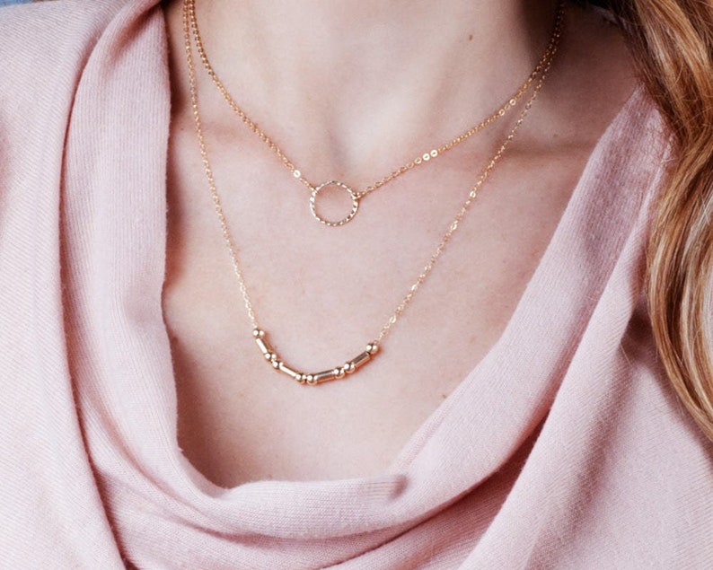 Puede incluir: Un collar de oro de varios niveles con un delicado colgante circular y una fila de peque&ntilde;as cuentas doradas.