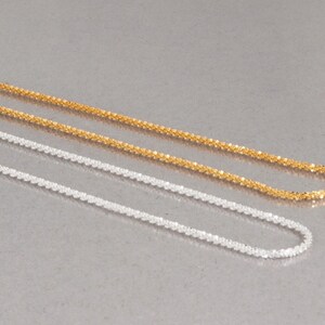 May include: Two delicate chains, one gold and one silver, lay side by side on a gray surface. Both chains are made of small, linked squares.
