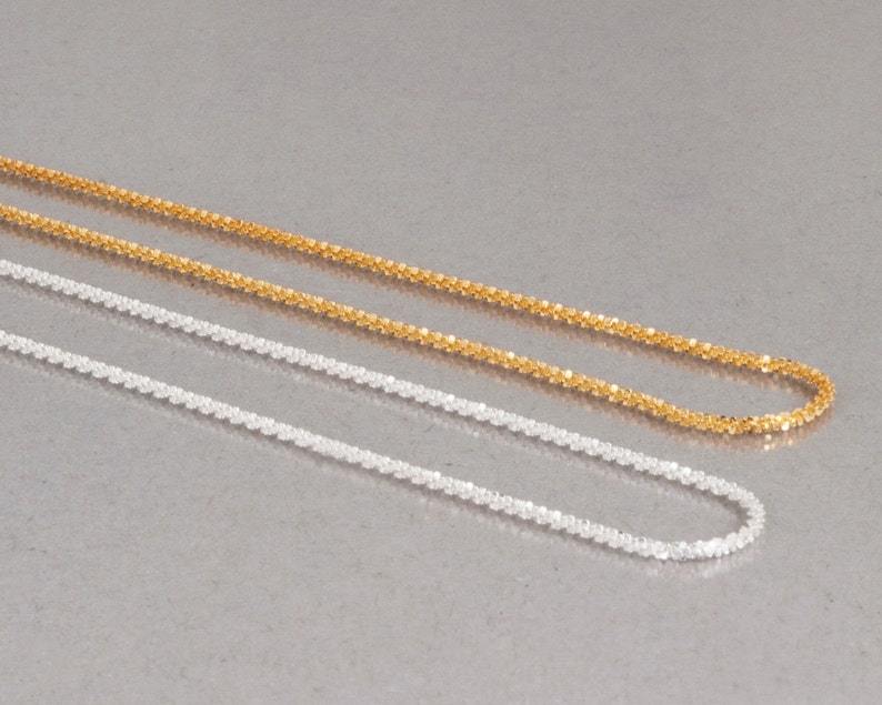 May include: Two delicate chains, one gold and one silver, lay side by side on a gray surface. Both chains are made of small, linked squares.