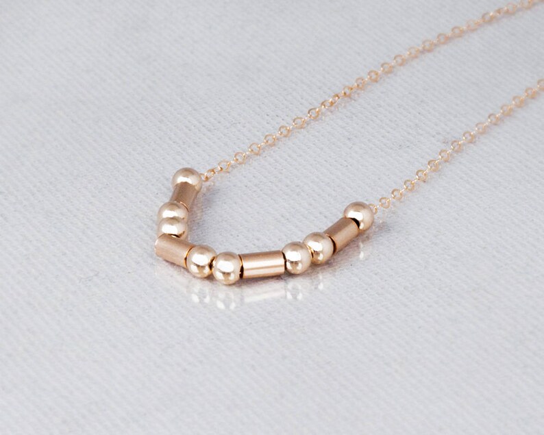 Puede incluir: Un collar de oro con una cadena delicada y una fila de cuentas y cilindros de oro alternados.
