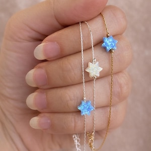 Puede incluir: Tres delicados collares con colgantes en forma de estrella. Un collar es dorado y los otros dos son plateados. Los colgantes de estrella son azules y blancos. Los collares se sostienen en una mano sobre un fondo rosa suave.