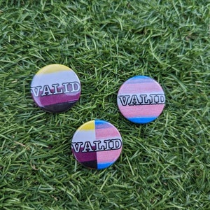 May include: Three round buttons with different pride flags and the word "VALID" printed in white on each button. The buttons are on a green grassy surface.