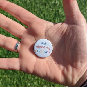 May include: A white button with the text "ask about my pronouns" in rainbow colours. The button is being held in a person's hand.