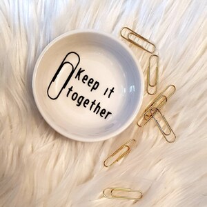 May include: A white ceramic dish with a gray ombre design and the text "Keep it together" with a paperclip graphic. The dish is surrounded by gold paperclips on a white fluffy surface.