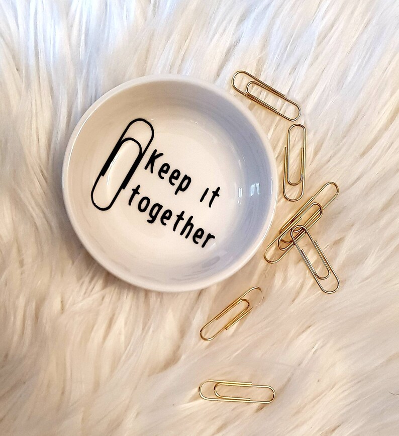 May include: A white ceramic dish with a gray ombre design and the text "Keep it together" with a paperclip graphic. The dish is surrounded by gold paperclips on a white fluffy surface.