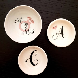 May include: Three white ceramic ring dishes with black lettering. The largest dish says "est. & mrs." with a pink diamond ring illustration. The medium dish says "A" with a pink heart. The smallest dish says "C" with two small hearts.