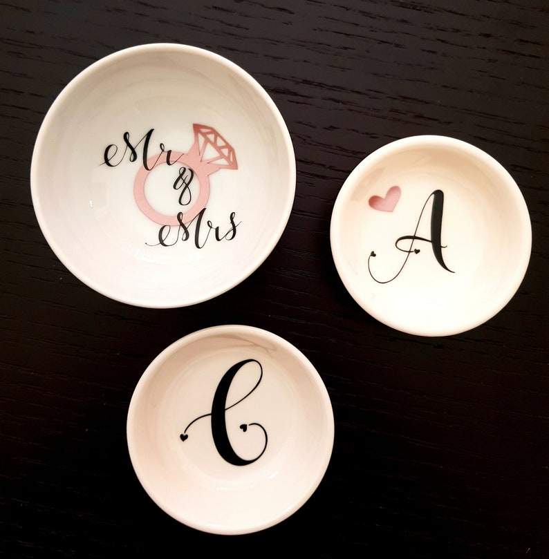 May include: Three white ceramic ring dishes with black lettering. The largest dish says "est. & mrs." with a pink diamond ring illustration. The medium dish says "A" with a pink heart. The smallest dish says "C" with two small hearts.