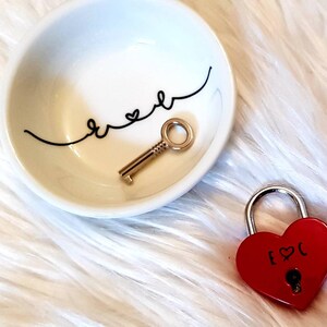 May include: A white ceramic dish with the word "forever" written in black cursive with a heart symbol. A gold key is inside the dish. A red heart-shaped padlock with a gold keyhole and the text "[heart]C" is on a white fluffy surface.