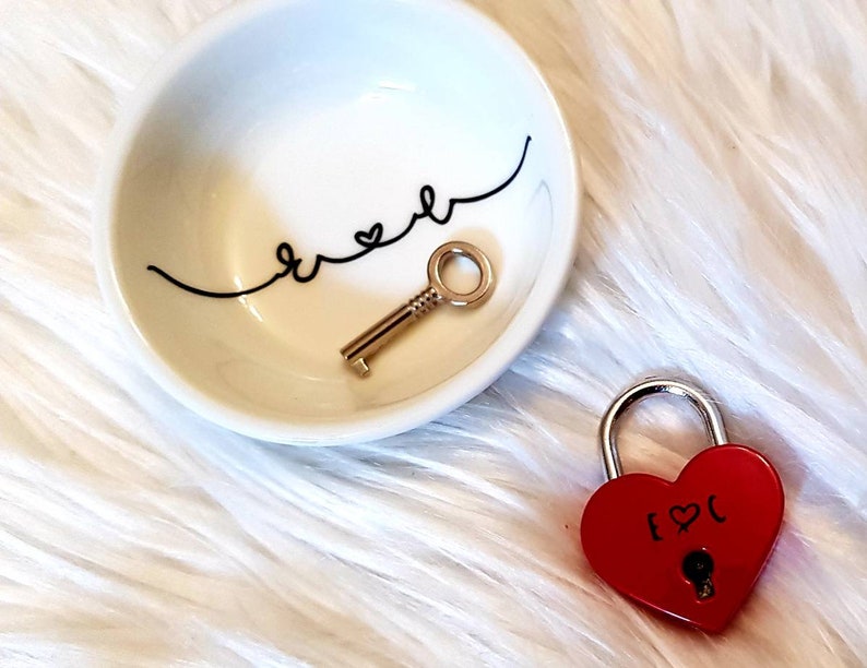 May include: A white ceramic dish with the word "forever" written in black cursive with a heart symbol. A gold key is inside the dish. A red heart-shaped padlock with a gold keyhole and the text "[heart]C" is on a white fluffy surface.
