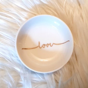 May include: A white ceramic ring dish with a gold inscription that reads "love".