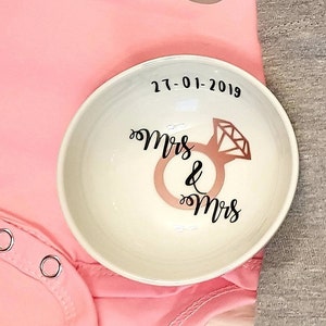 May include: White ceramic ring dish with the text "Mrs & Mrs" and a diamond ring design. The date "27-01-2019" is written above the ring design.