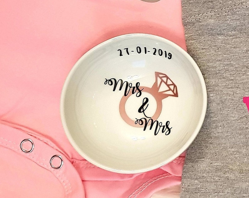 May include: White ceramic ring dish with the text "Mrs & Mrs" and a diamond ring design. The date "27-01-2019" is written above the ring design.