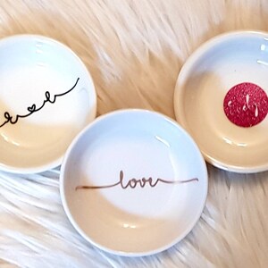 May include: Three small white ceramic dishes with decorative designs. One dish has the word "sweet" written in black cursive, another has the word "love" written in gold cursive, and the third has a red glitter circle with the word "bride" written in white.