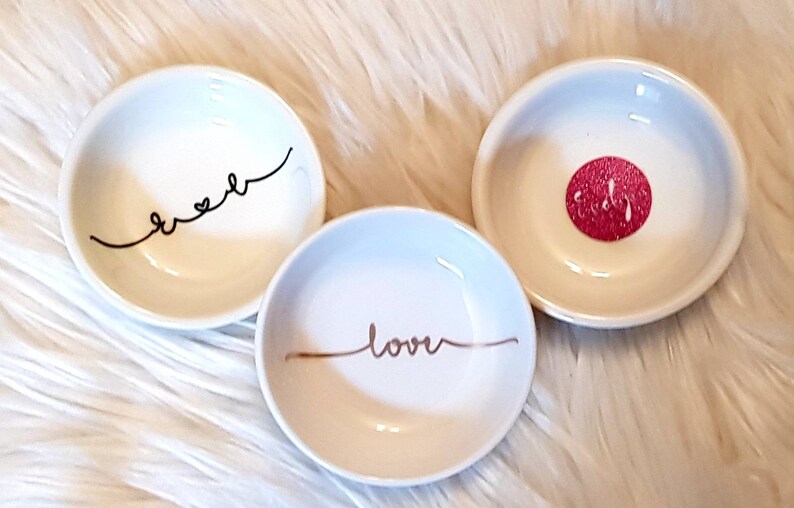 May include: Three small white ceramic dishes with decorative designs. One dish has the word "sweet" written in black cursive, another has the word "love" written in gold cursive, and the third has a red glitter circle with the word "bride" written in white.