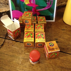 May include: A display of small, gold boxes of "BASKAR PERFUMERS" with parrot illustrations, a red-topped jar, and a framed floral print. The boxes are arranged on a wooden surface. The boxes are labelled "HAND MADE PRODUCT".