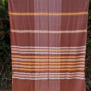 Peut inclure: Textile tissé marron avec des rayures verticales et des bandes horizontales blanches, orange et jaunes. Le textile a un bord frangé et est exposé à l'extérieur, suspendu devant un feuillage vert.