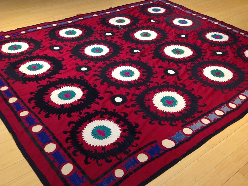 K&ouml;nnte beinhalten: Ein roter Stoffteppich mit einem sich wiederholenden Muster aus schwarzen, wei&szlig;en und gr&uuml;nen Kreisen. Die Kreise haben ein stilisiertes Blumenmuster um sie herum.