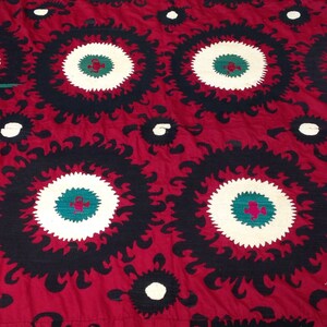 K&ouml;nnte beinhalten: Ein roter Stoff mit einem sich wiederholenden Muster aus schwarzen und wei&szlig;en Kreisen mit gr&uuml;nen und roten Akzenten. Der Stoff hat ein traditionelles Design.