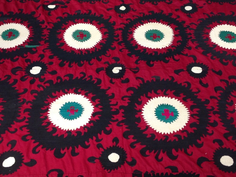 K&ouml;nnte beinhalten: Ein roter Stoff mit einem sich wiederholenden Muster aus schwarzen und wei&szlig;en Kreisen mit gr&uuml;nen und roten Akzenten. Der Stoff hat ein traditionelles Design.