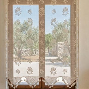 Puede incluir: Cortinas de ventana de encaje beige con un estampado floral de rosas. Las cortinas cuelgan de un marco de ventana de madera, con una vista de árboles y una pared de piedra visible a través de la ventana. El borde inferior tiene borlas.