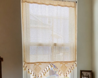 May include: Beige window curtain with a scalloped top and a decorative bottom edge. The bottom edge features a geometric pattern with hanging tassels. The curtain is made of a woven material, allowing light to filter through.