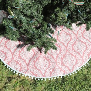 May include: A pink and white Christmas tree skirt with a damask pattern and a white pom-pom trim.