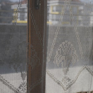 May include: A white crocheted curtain with a delicate pattern. The curtain is hanging in a window and the view outside is blurred.