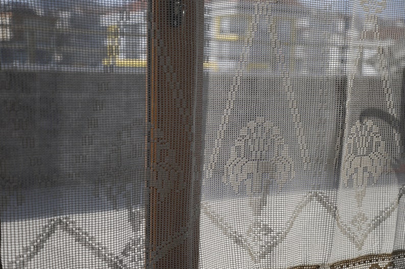 May include: A white crocheted curtain with a delicate pattern. The curtain is hanging in a window and the view outside is blurred.