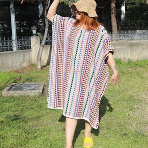 May include: A loose-fitting, knee-length crochet tunic featuring a vertical pattern of multicoloured dots in shades of blue, red, green, and yellow on a white background, paired with a tan woven straw bucket hat and bright yellow slide sandals.