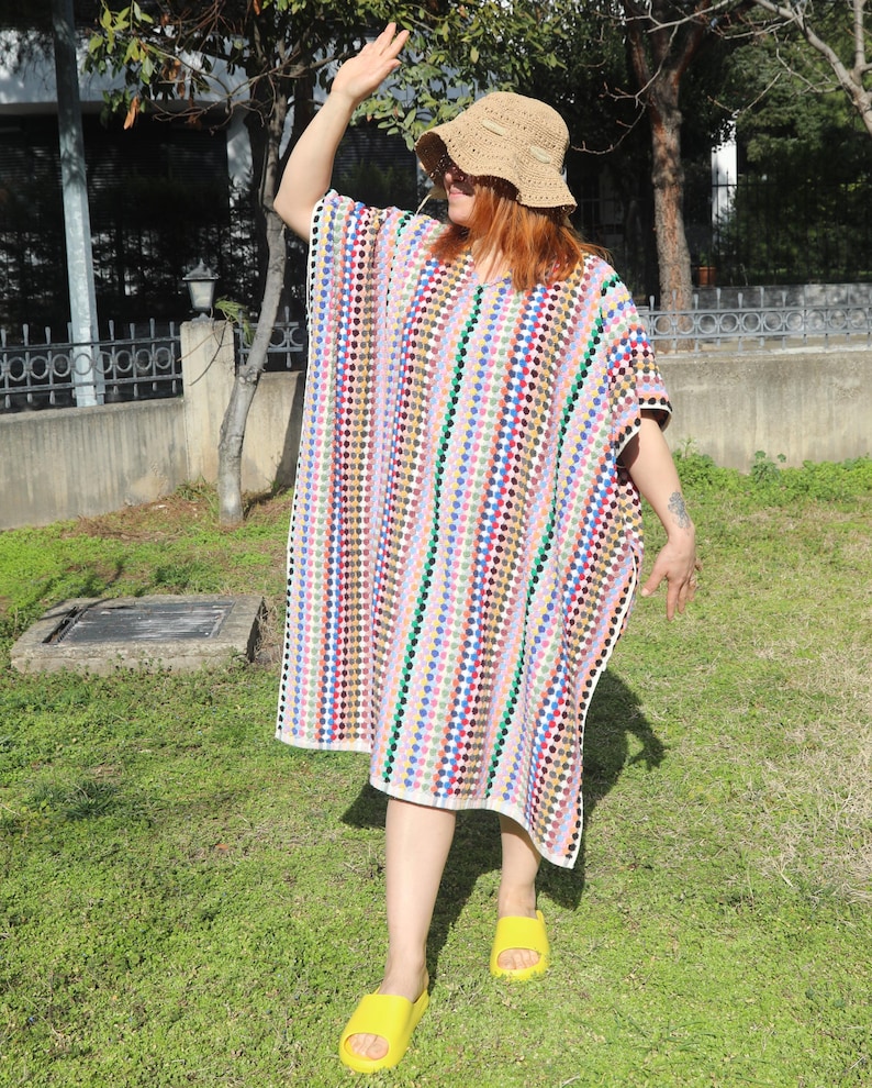 May include: A loose-fitting, knee-length crochet tunic featuring a vertical pattern of multicoloured dots in shades of blue, red, green, and yellow on a white background, paired with a tan woven straw bucket hat and bright yellow slide sandals.