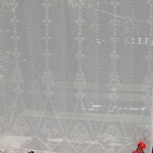 May include: White crocheted curtain with a scalloped bottom and tassels. The curtain has a delicate floral pattern and is hung in front of a window.
