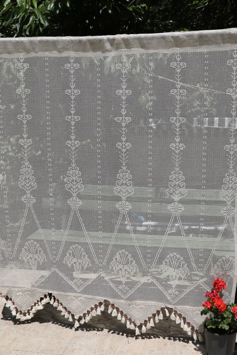 May include: White crocheted curtain with a scalloped bottom and tassels. The curtain has a delicate floral pattern and is hung in front of a window.