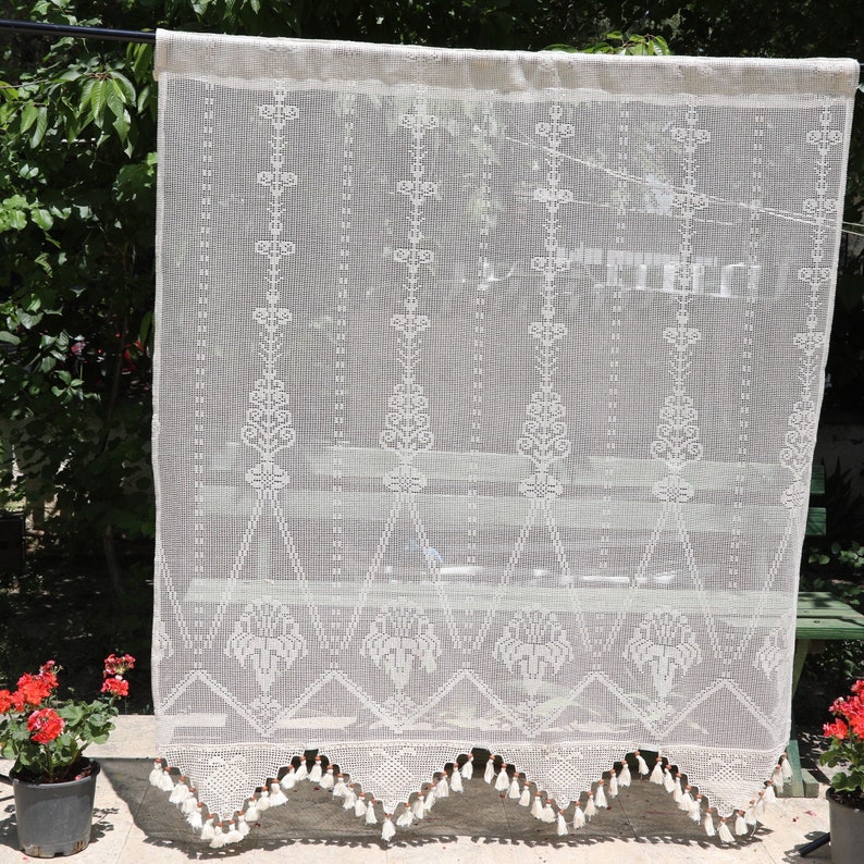 May include: White crocheted curtain with a scalloped bottom and fringe trim. The curtain has a delicate floral pattern.