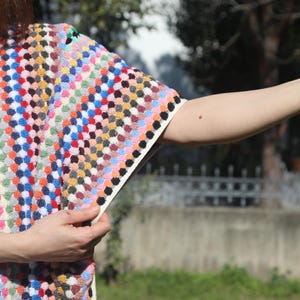 May include: A short-sleeved top featuring a dense, multi-coloured textured pattern of small, raised dots in shades of red, blue, yellow, green, and white. The garment has a V-neckline and a cream-coloured trim along the sleeve edge and side seam.