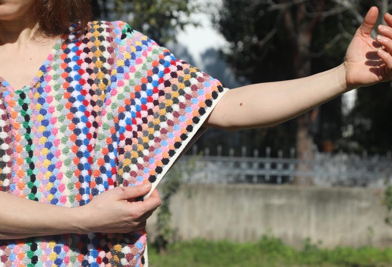 May include: A short-sleeved top featuring a dense, multi-coloured textured pattern of small, raised dots in shades of red, blue, yellow, green, and white. The garment has a V-neckline and a cream-coloured trim along the sleeve edge and side seam.