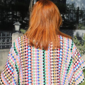 May include: A crochet poncho featuring a vibrant, multi-coloured pattern of small, raised circular stitches in shades of red, blue, yellow, green, and black. The garment has a wide, loose-fitting silhouette with a textured, knit-like appearance.