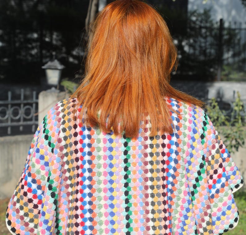 May include: A crochet poncho featuring a vibrant, multi-coloured pattern of small, raised circular stitches in shades of red, blue, yellow, green, and black. The garment has a wide, loose-fitting silhouette with a textured, knit-like appearance.