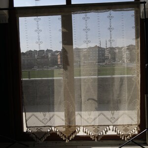 May include: A window with a sheer lace curtain, showing a view of a city skyline with buildings and a mosque in the distance.
