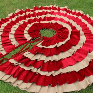 May include: A red and beige ruffled Christmas tree skirt with a circular shape. The skirt is made of burlap and velvet and has a tied closure.