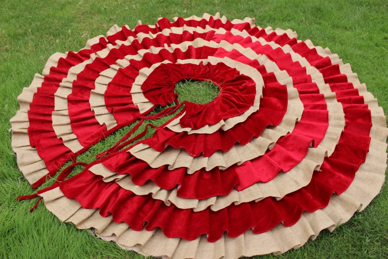 May include: A red and beige ruffled Christmas tree skirt with a circular shape. The skirt is made of burlap and velvet and has a tied closure.